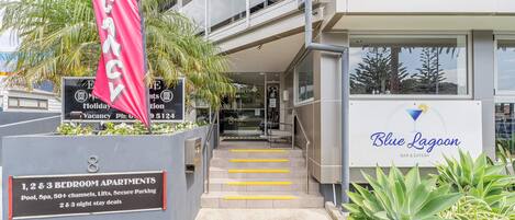A building with a sign for 'Blue Lagoon' bar and eatery, and a sign for 'Vacancy' with a phone number. There are 1, 2, and 3 bedroom apartments available, with amenities like pool, spa, 50+ channels, lifts, and secure parking.