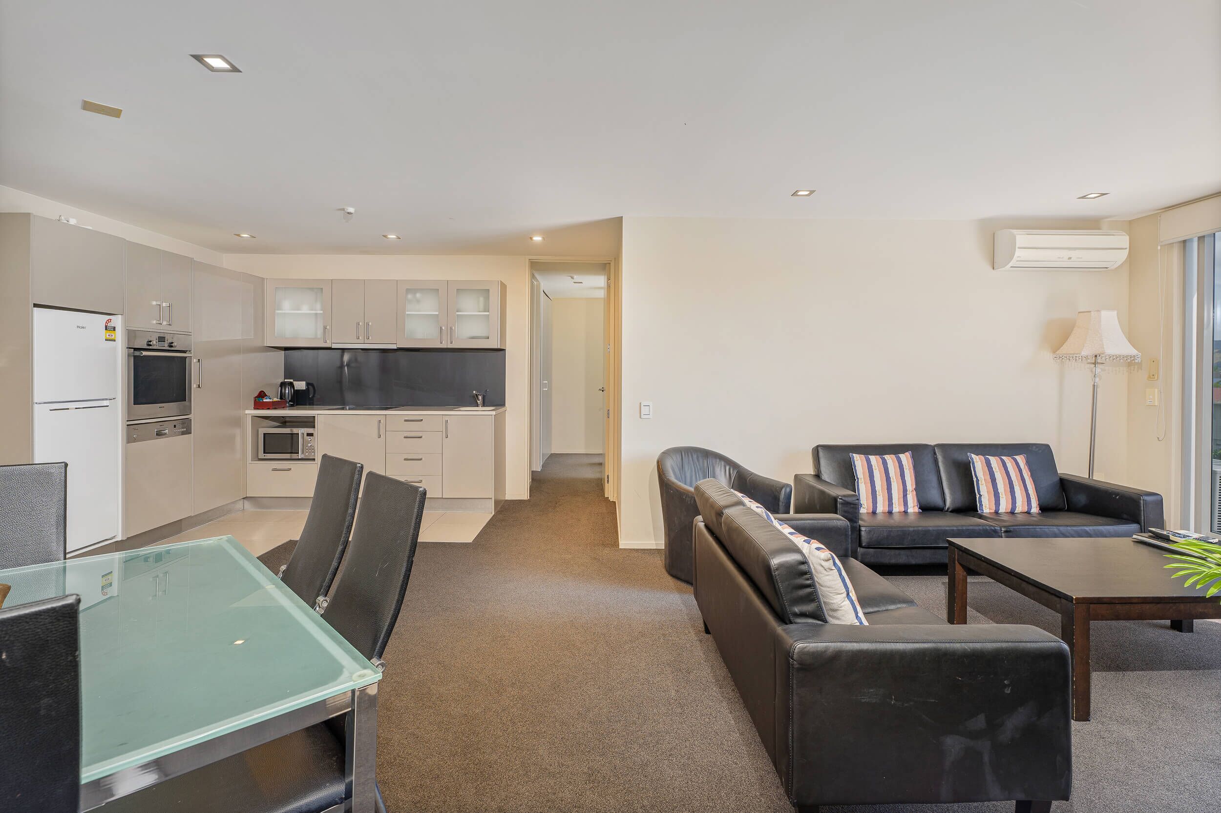 A modern living room with a glass-top dining table, black leather sofas, and a black coffee table. There is a kitchen area with white cabinets and a microwave.
