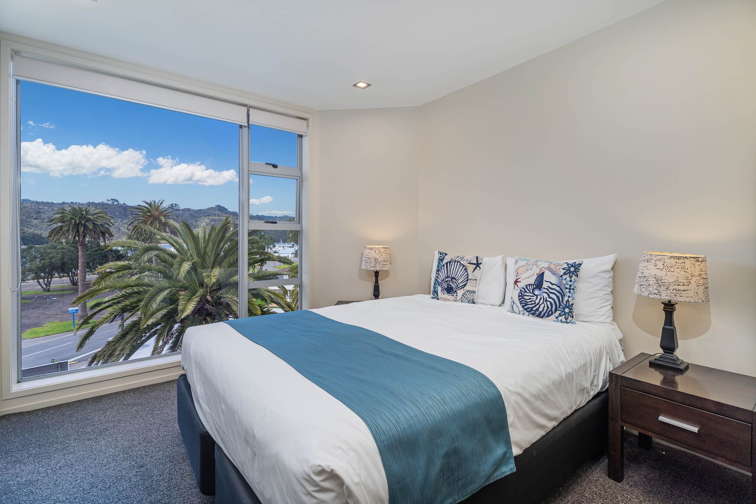 A bedroom with a large window, a bed with white and teal bedding, two bedside lamps, and decorative pillows.