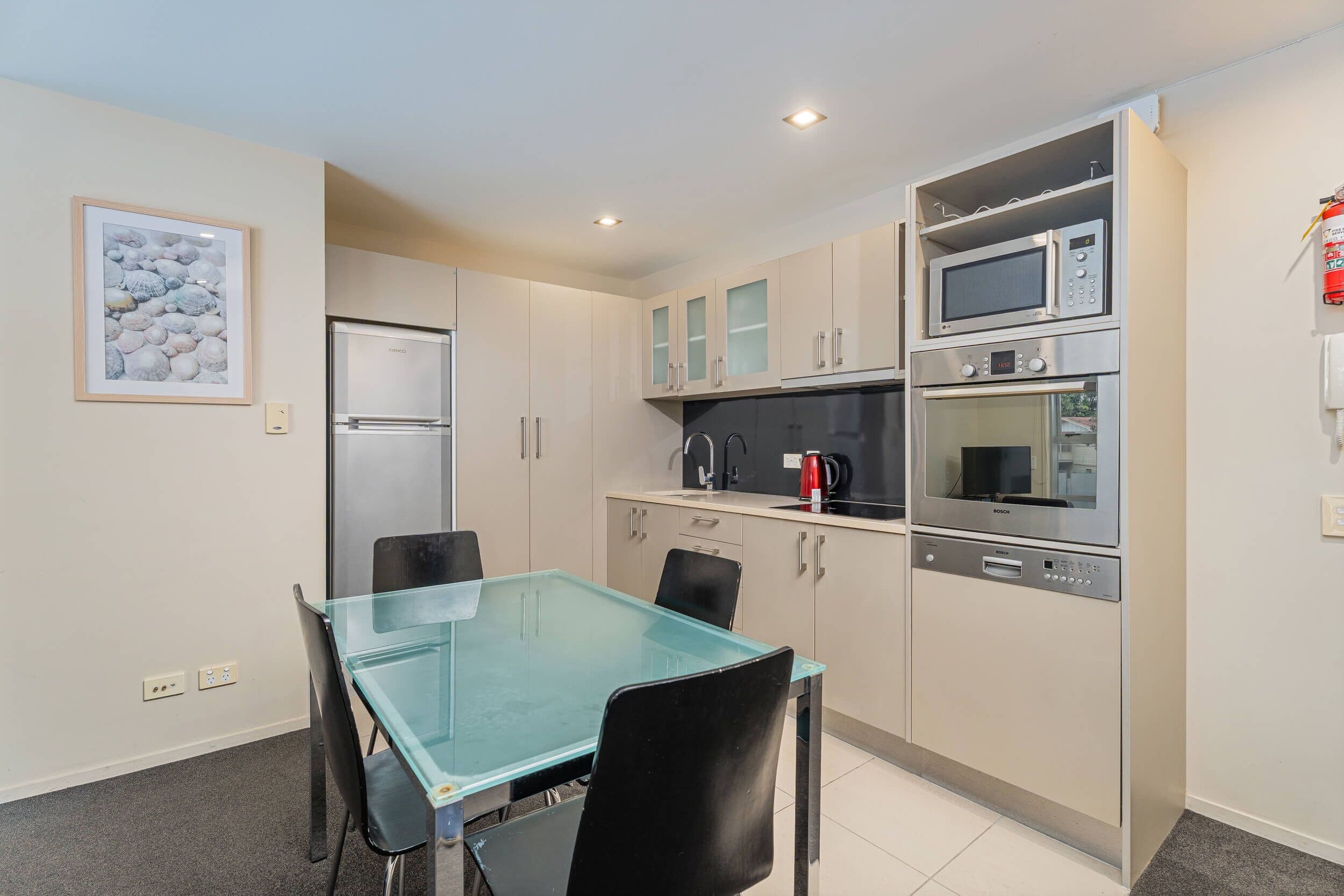 Cocina compacta con refrigerador, microondas, horno y lavavajillas. Hay una mesa de vidrio con sillas y un cuadro en la pared.