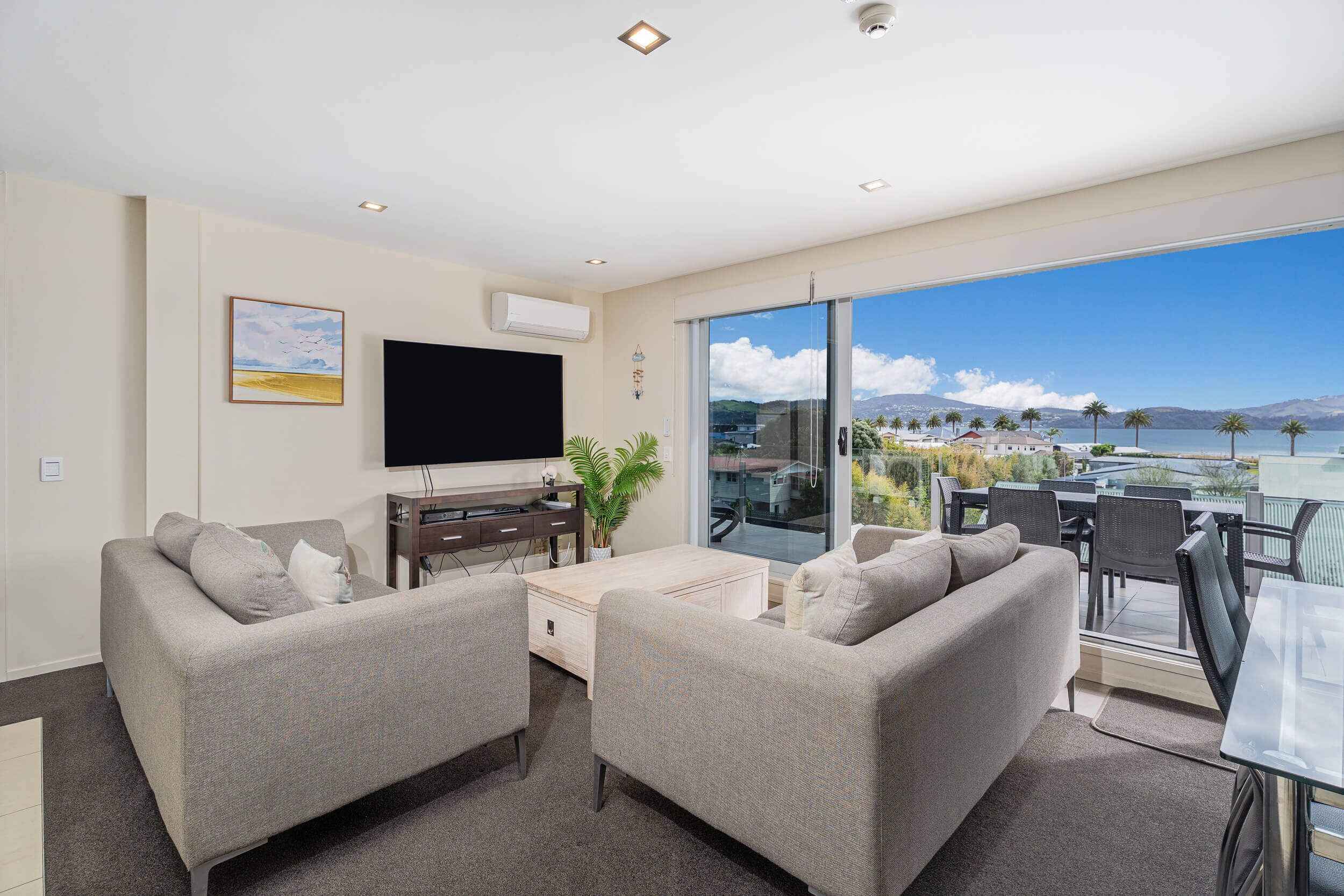 A modern living room with a flat-screen TV, two sofas, a coffee table, and a balcony with a view of the sea.
