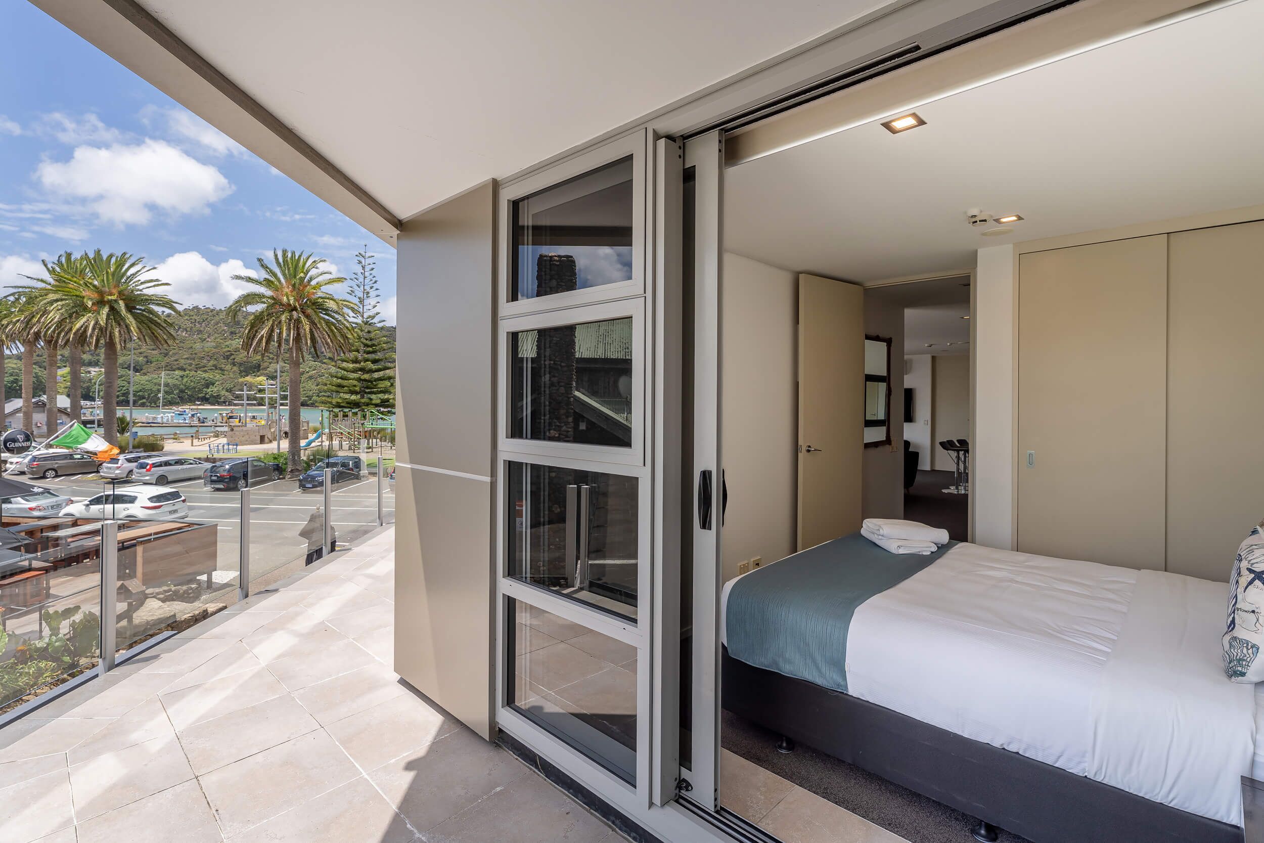 Un balcón con puerta de vidrio que da a un dormitorio con cama y vista a palmeras y un estacionamiento.