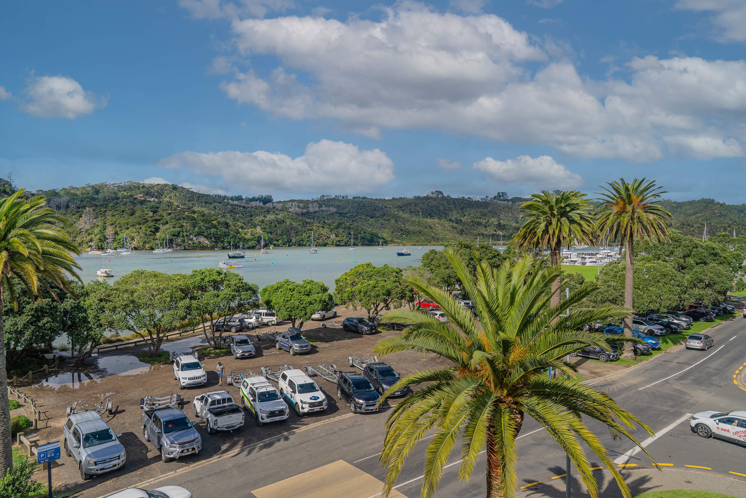 A coastal parking area with vehicles, a road, and a view of boats on the water.