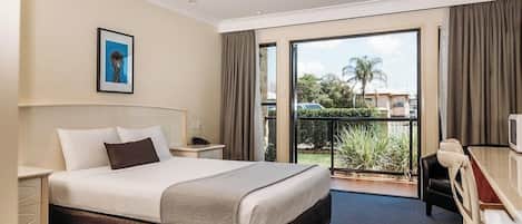 Habitación de hotel con una cama grande, un escritorio, una silla y un balcón con vistas a palmeras y edificios.