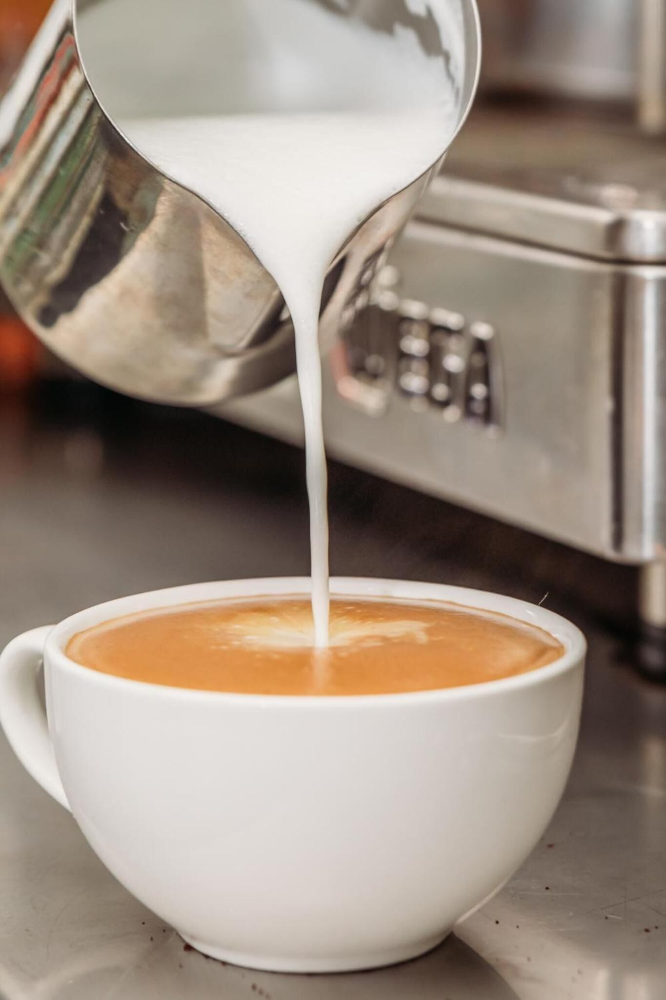 Una taza blanca de café con leche siendo servida desde una jarra de metal.