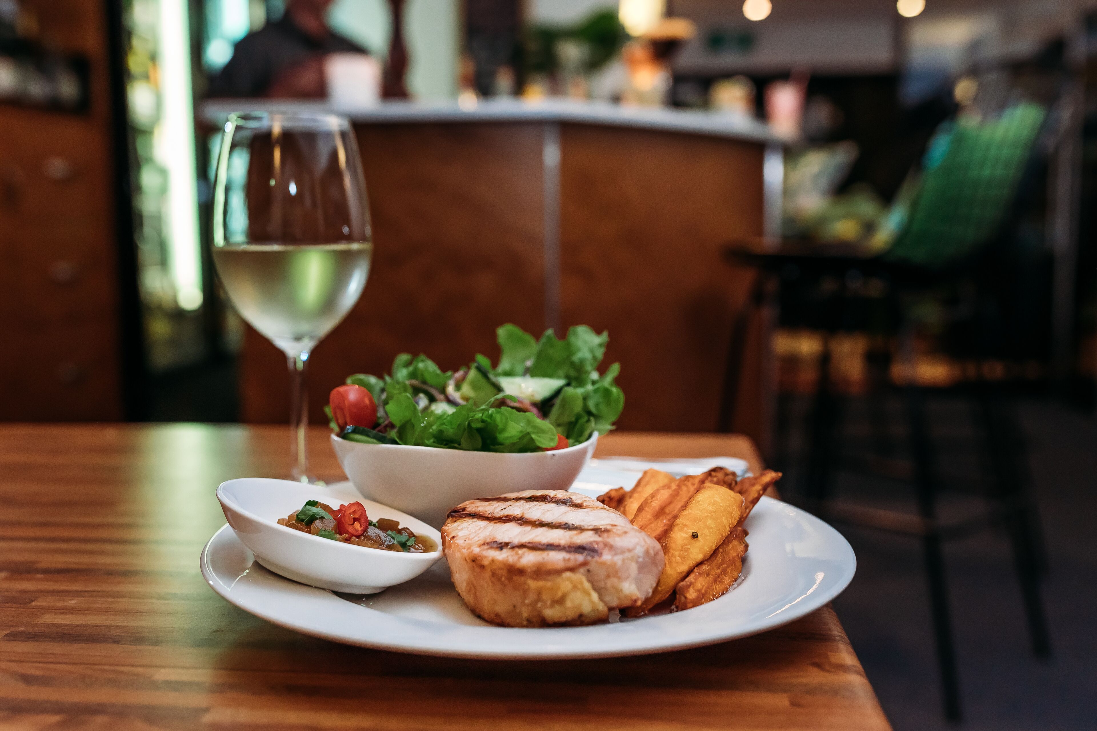 A plate with grilled meat, a salad, and a glass of white wine on a wooden table.