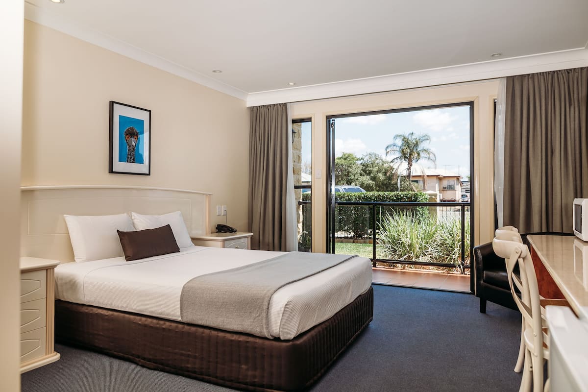 Une chambre d’hôtel avec un grand lit, un bureau, une chaise, une télévision et un balcon offrant une vue sur les palmiers et les immeubles.
