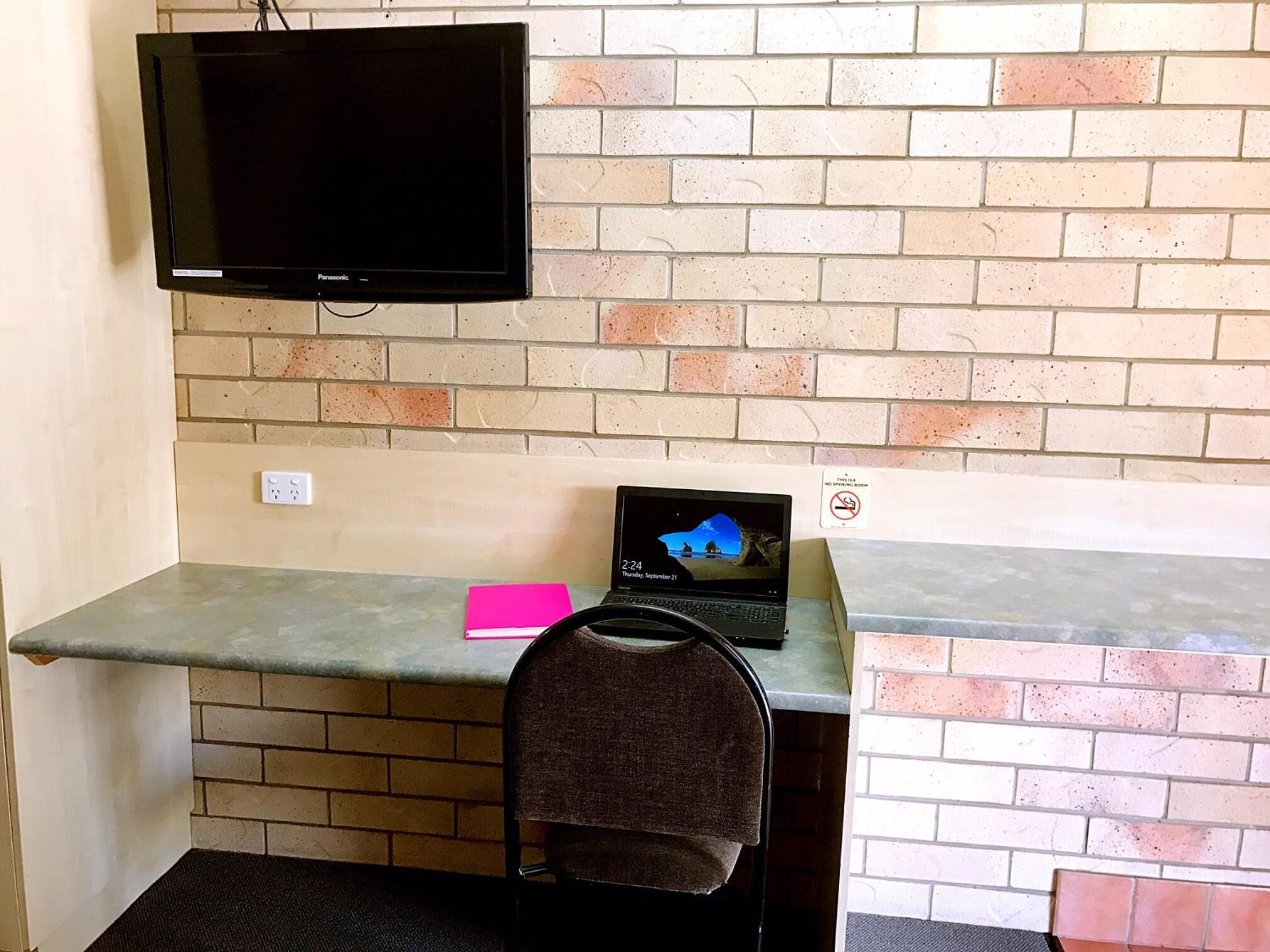 A room with a brick wall, a flat-screen TV mounted above a desk, a laptop, a pink notepad, and a chair with a towel.