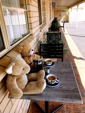 A table with a teddy bear, a coffee pot, and two cups of coffee on a patio with a brick wall and a window.