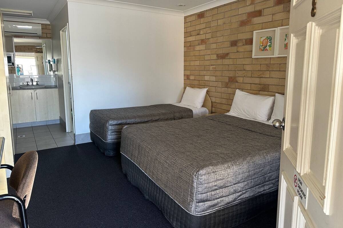 A hotel room with two beds, a ceiling fan, a brick wall, and a kitchen area visible through an open door.