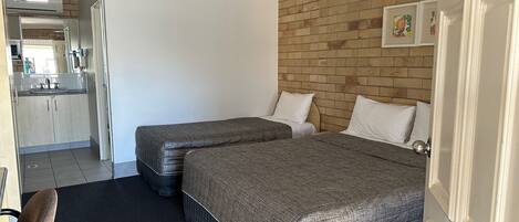Habitación de hotel con dos camas, ventilador de techo, pared de ladrillo y una cocina visible a través de una puerta abierta.