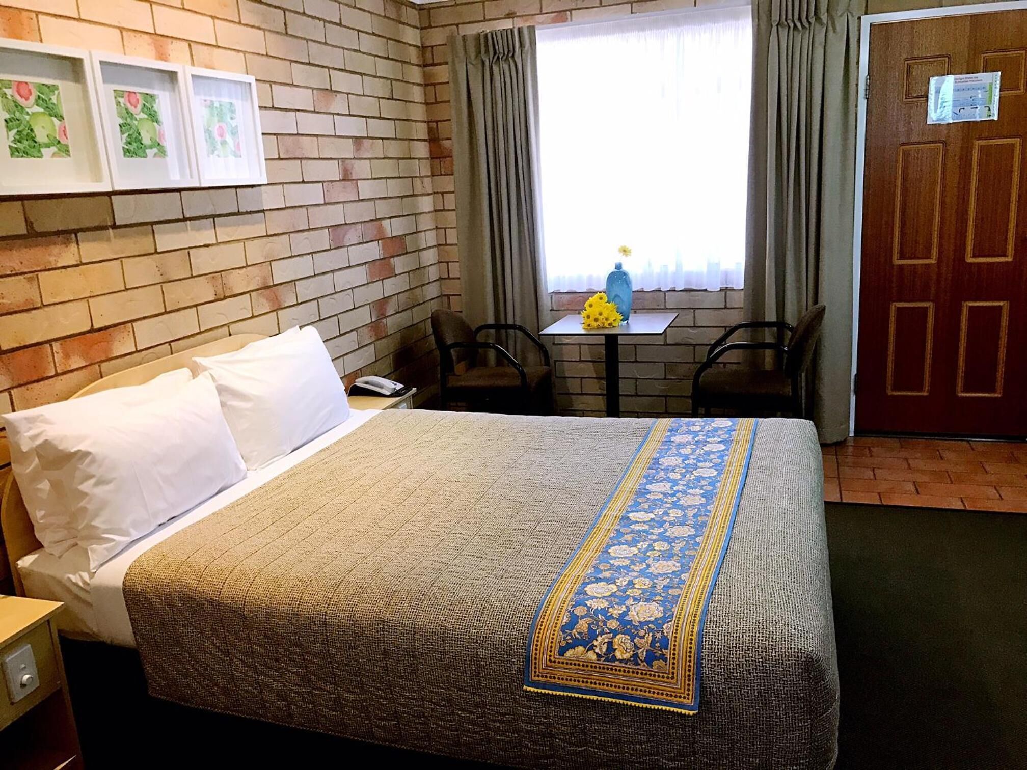 A hotel room with a bed, a small table with a vase, and a chair.