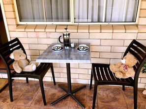 A small outdoor table with two chairs, each with a teddy bear. There is a coffee pot and two cups on the table.
