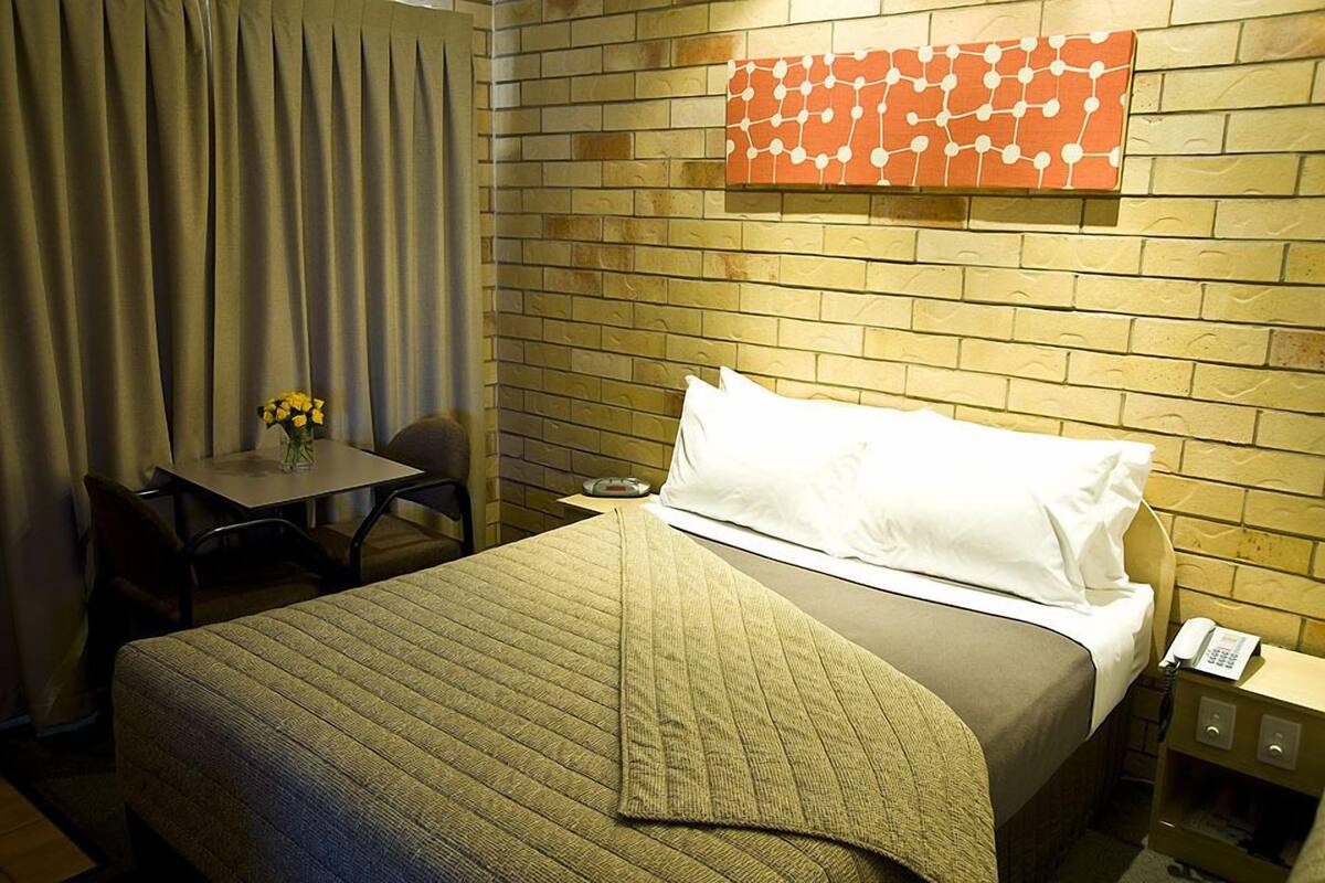 A hotel room with a bed, a small table with a vase of flowers, a chair, and a wall-mounted telephone.