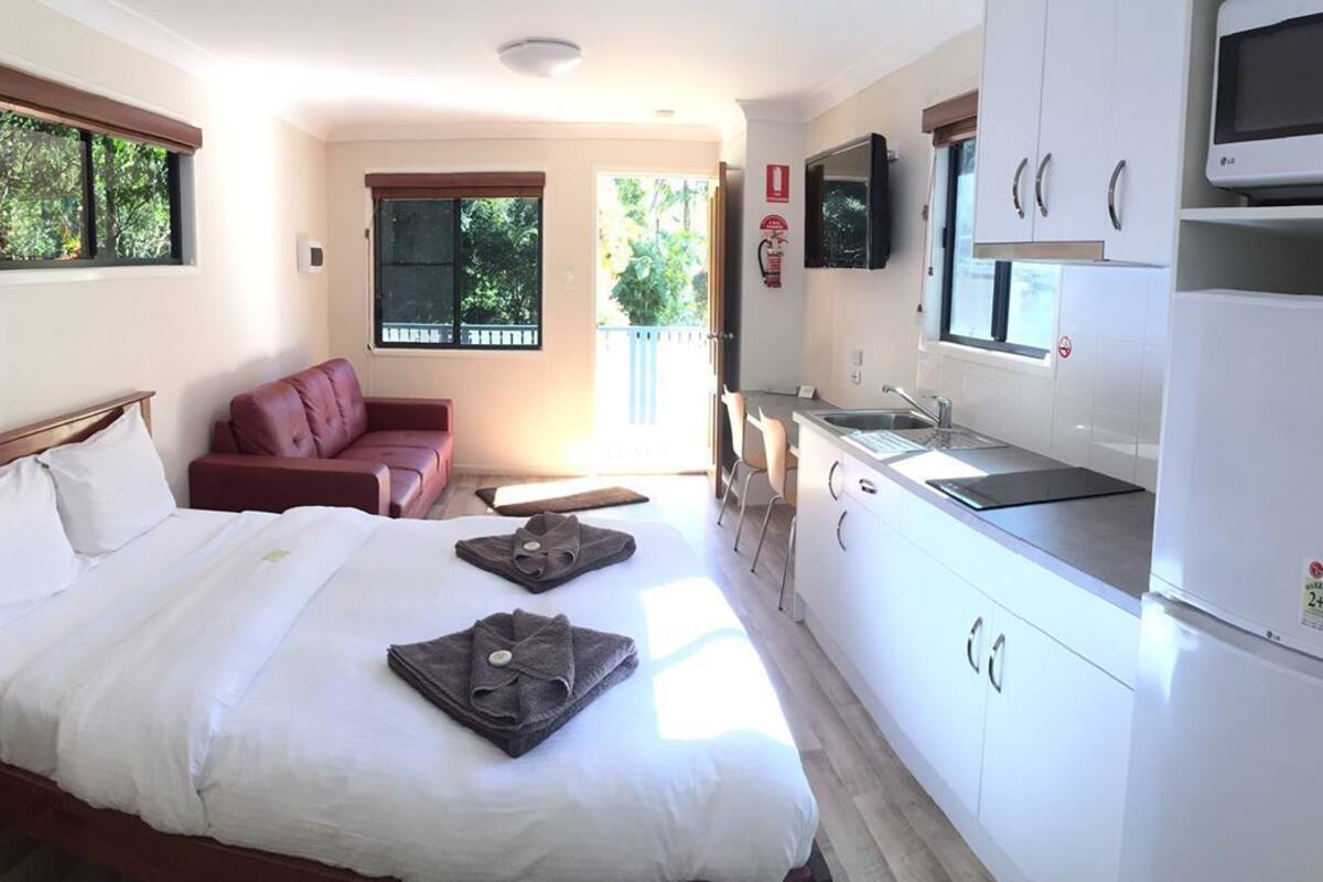 A hotel room with a bed, a desk, a chair, a kitchenette, and a window with blinds.