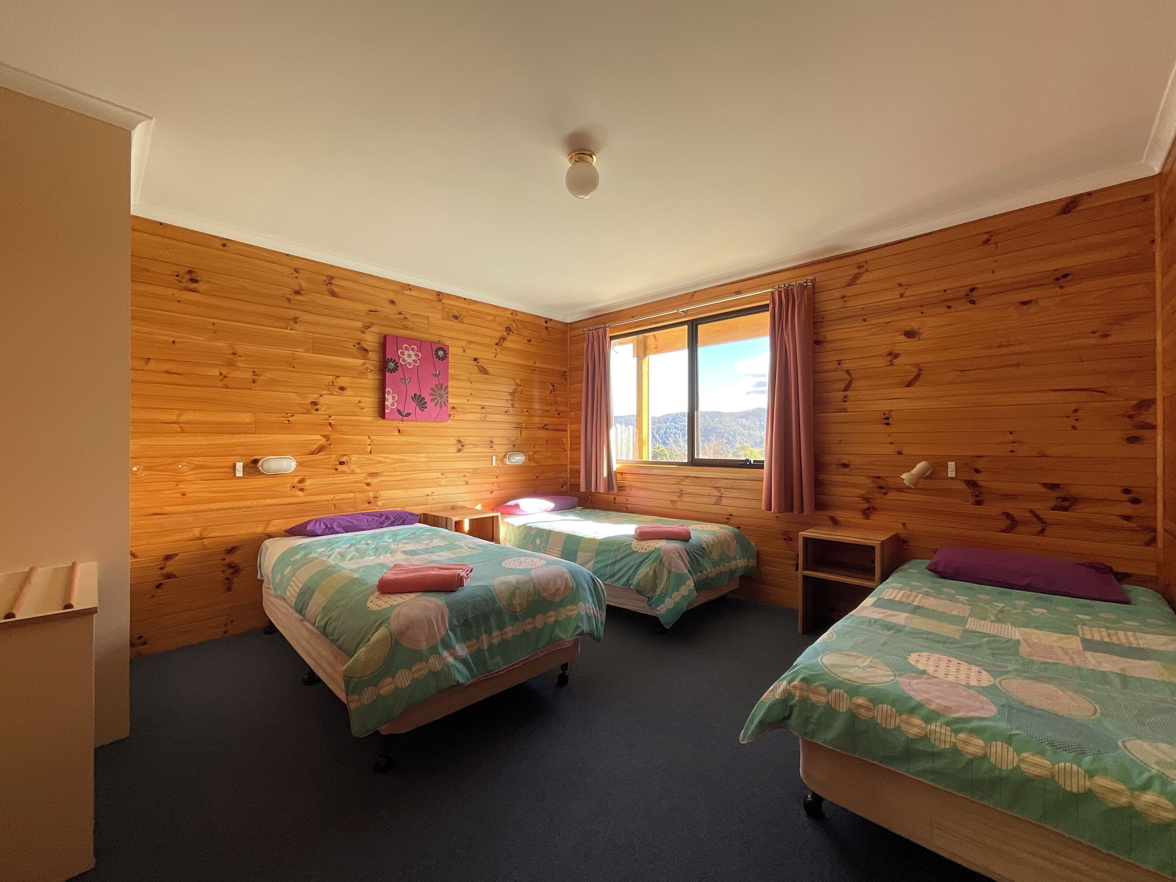 A room with wooden walls, two beds with green bedding, a window with curtains, and a small table.