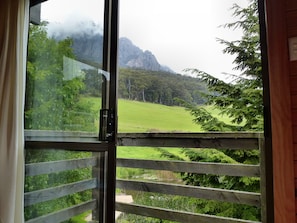 A view from a room with a wooden fence and a glass door leading to a green landscape with mountains in the background.