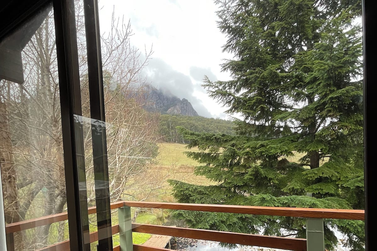 A view from a balcony with a wooden railing, overlooking a pond and a forested area with mountains in the background.