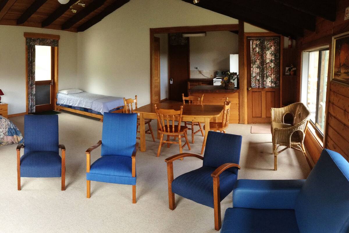 A cozy room with a bed, two blue chairs, a wooden dining table, and a kitchen area.