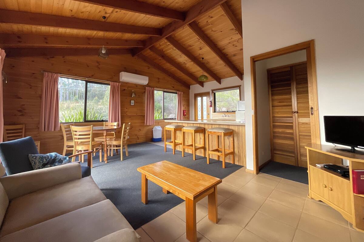 A wooden cabin interior with a sofa, dining table, chairs, a kitchen area, and a TV.