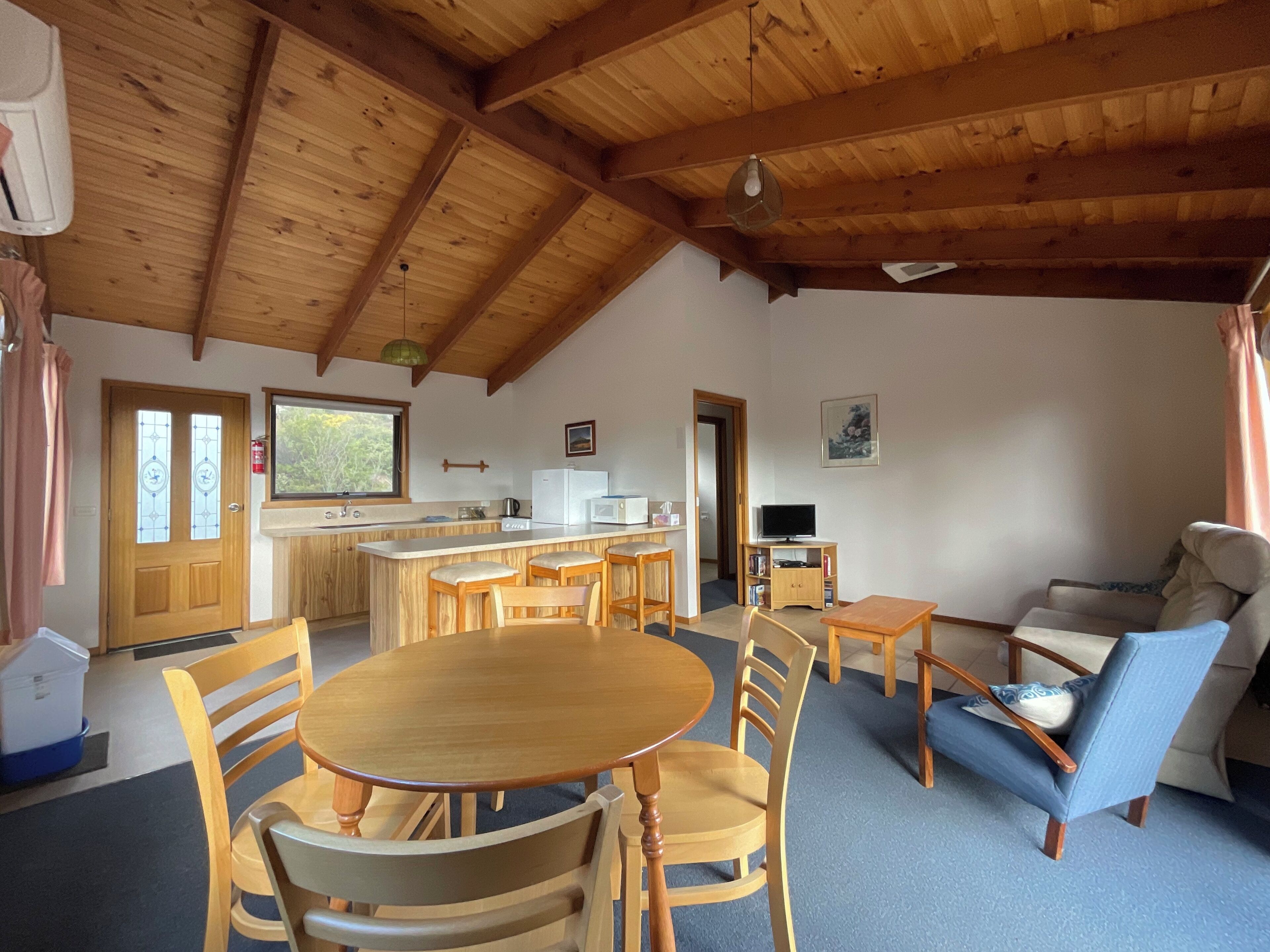 A spacious living area with a wooden ceiling, a dining table with chairs, a sofa, and a television.