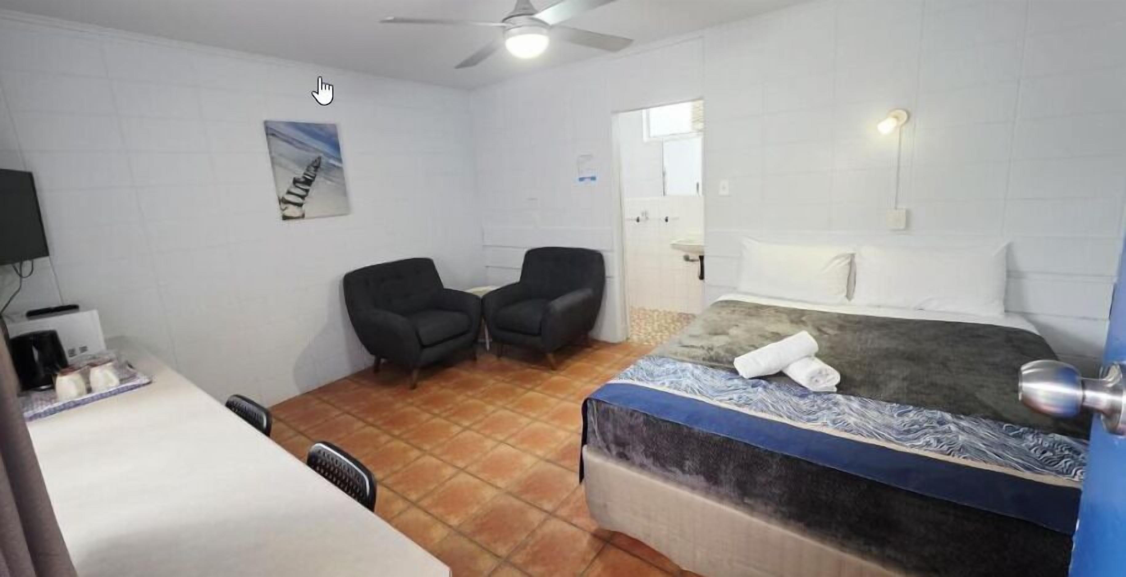 A hotel room with a bed, two chairs, a desk, a television, and a ceiling fan.