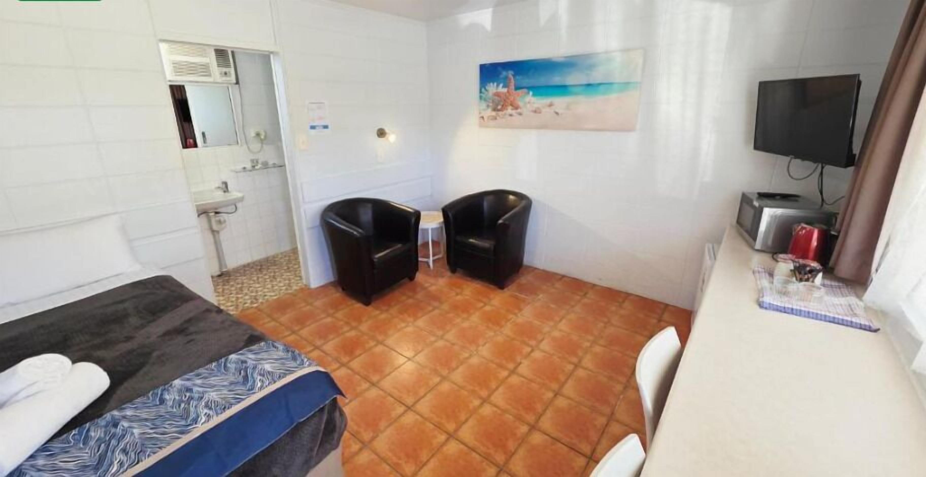 Una habitación con una cama, dos sillas, un escritorio, un televisor y un cuadro de una playa.
