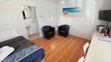 Una habitación con una cama, dos sillas, un escritorio, un televisor y un cuadro de una playa.