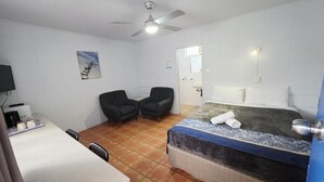 Habitación de hotel con cama, dos sillas, escritorio con cafetera, ventilador de techo y televisor montado en la pared.