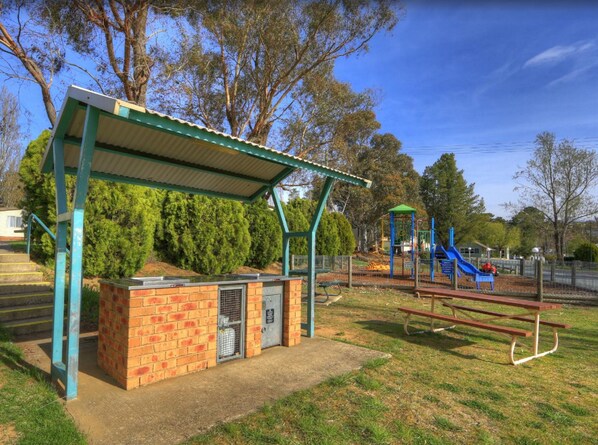 A park with a covered picnic area, a playground, and a brick structure with a ventilation unit. - NRMA Jindabyne Holiday Park (Jindabyne)