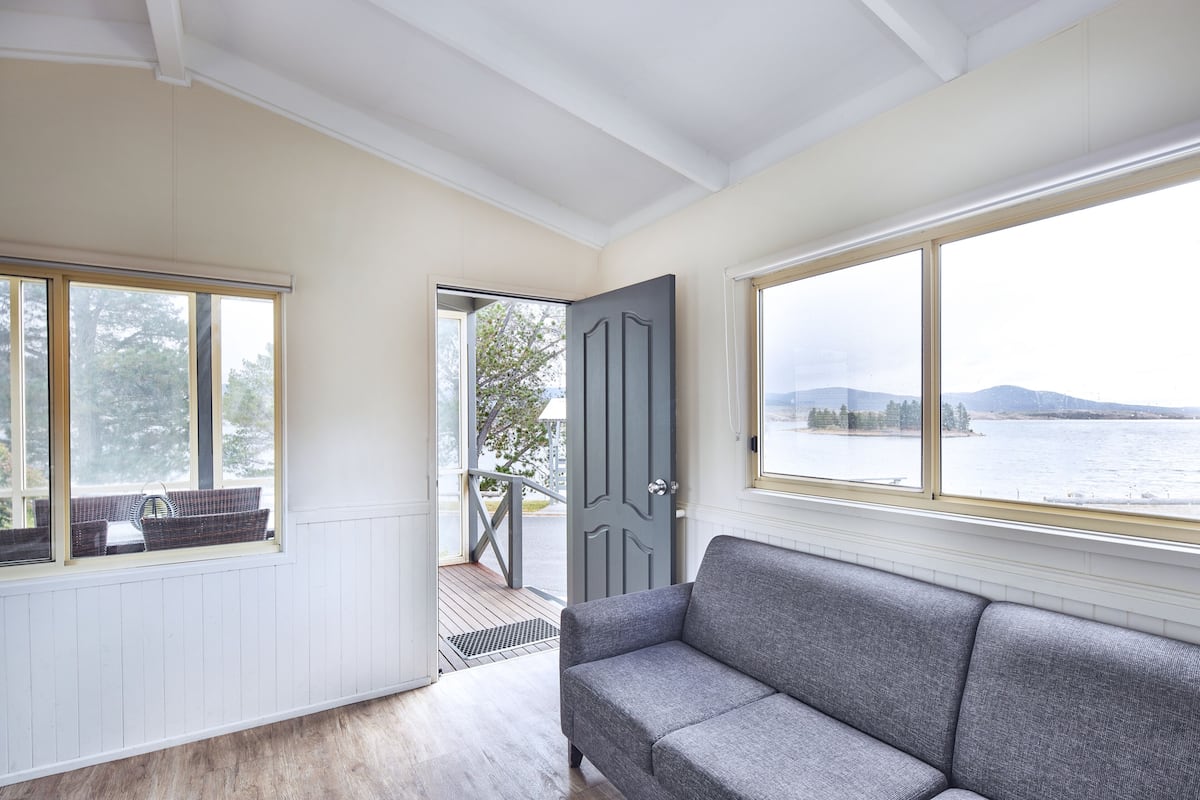 Una habitación con un sofá gris, un ventanal con vistas a un lago y una puerta que da al exterior.