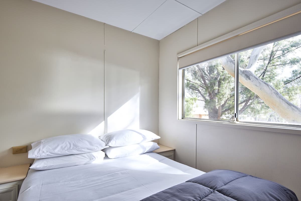 Un dormitorio con cama, mesita de noche, ventana con vistas a árboles y toma de corriente.