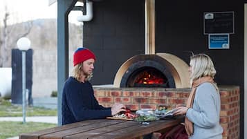 Un horno de ladrillo al aire libre con fuego encendido, una mesa de madera con comida y una pizarra con escritura.