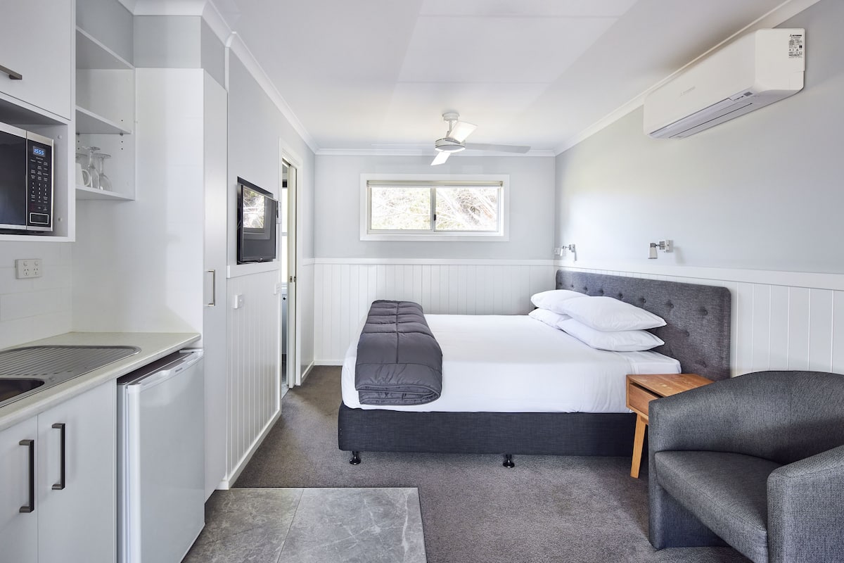 Habitación pequeña con cama, mesita de noche, una silla, ventilador de techo y ventana.