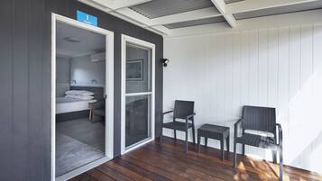 A room with a wooden floor, two chairs, a small table, and a door leading to a bathroom with a bed and a toilet.