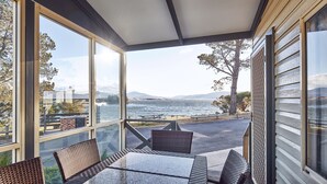 Eine überdachte Terrasse mit Tisch und Stühlen, große Fenster mit Blick auf Wasser und Berge.