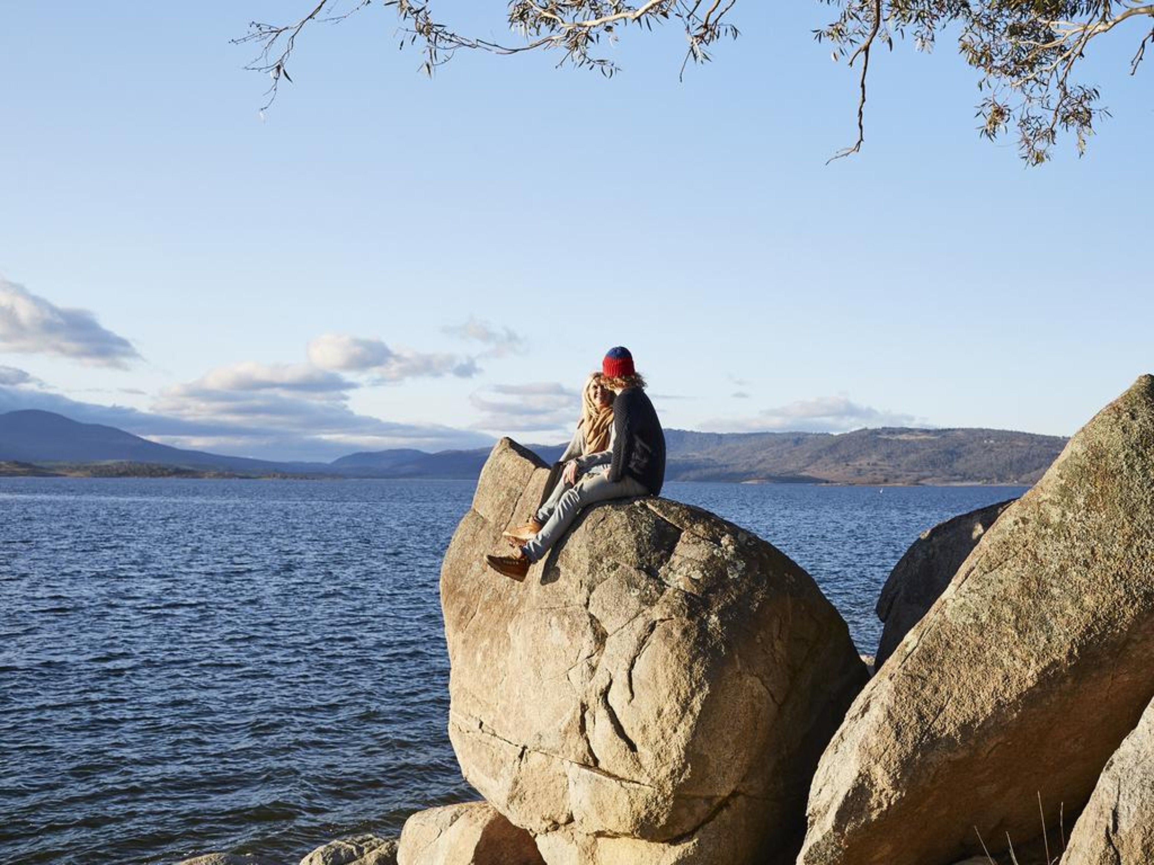 En person sitter på en stor sten vid en sjö eller ett annat vattendrag, med berg i bakgrunden.