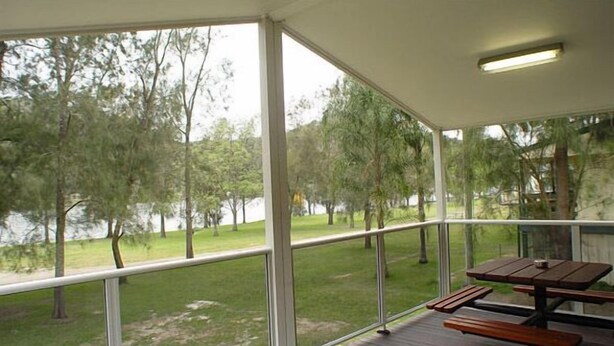 Un espacio al aire libre techado con una mesa de picnic de madera y bancos, con vistas a una zona verde con árboles y un cuerpo de agua.