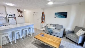 Una sala de estar moderna con área de cocina, muebles blancos, una mesa de centro de madera y un cuadro en la pared.