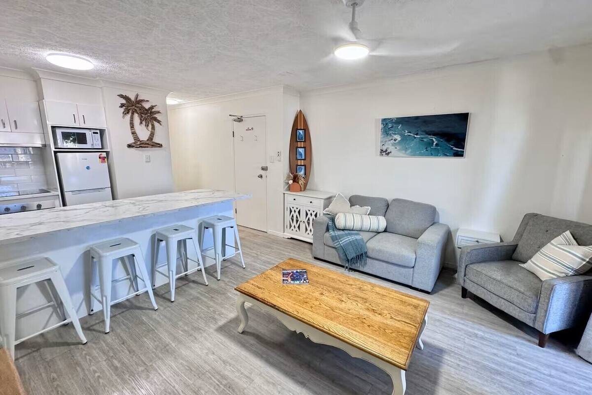 A modern living room with a kitchen area, white furniture, a wooden coffee table, and a painting on the wall.