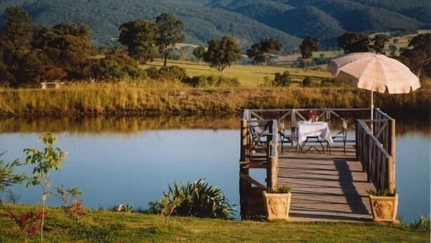 A lakeside wooden dock with a table and chairs, surrounded by lush greenery and hills in the background. - Wombat Hills Cottages (Barwite)