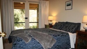 A bedroom with a large bed, two bedside lamps, a window with curtains, and a framed picture on the wall. - Wombat Hills Cottages (Barwite)