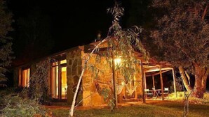 A stone building with a glass door and windows, surrounded by trees and illuminated by warm lighting. - Wombat Hills Cottages (Barwite)