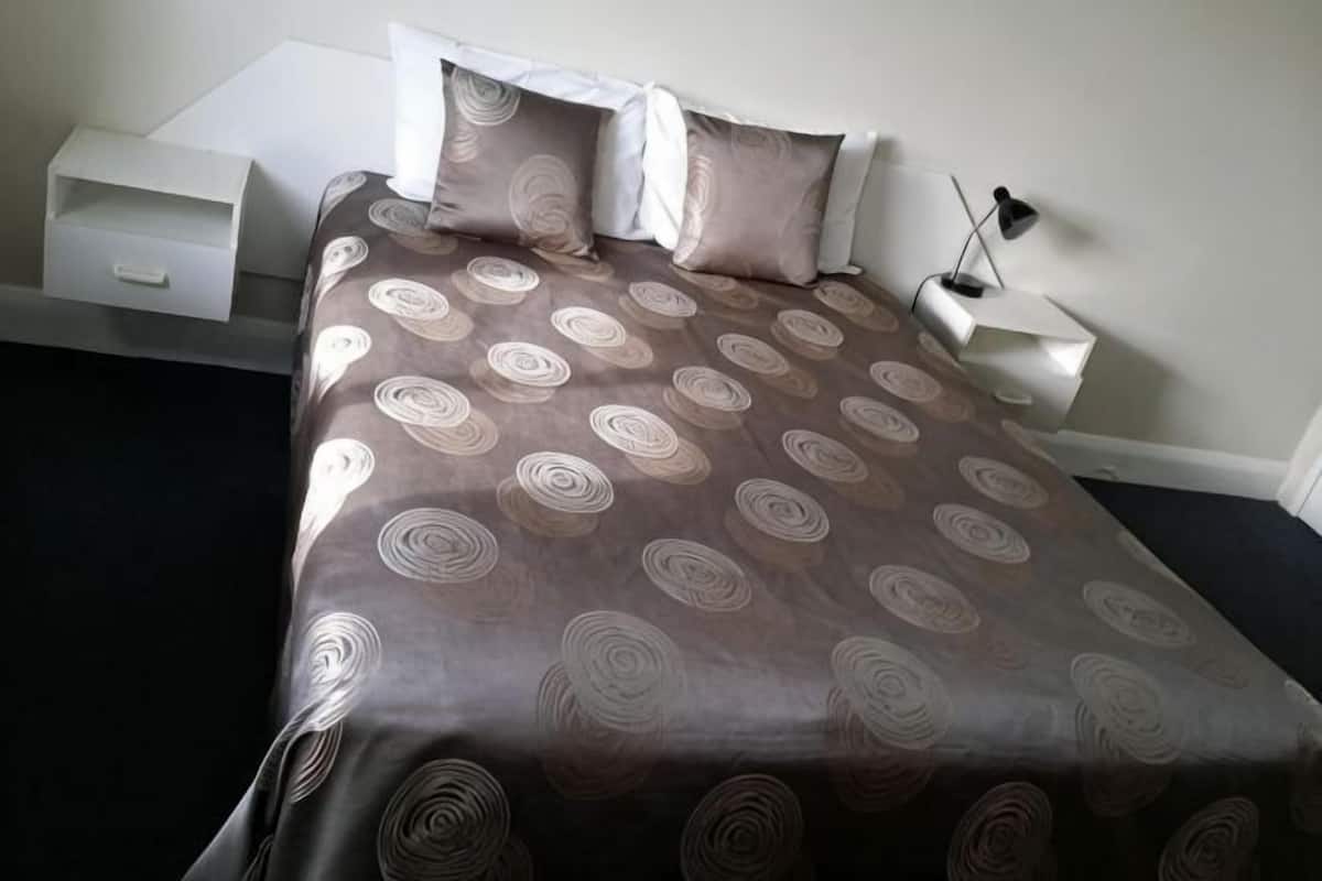 Un lit bien fait, recouvert d’une couvre-lit à motifs, une table de chevet avec une lampe et une petite table de nuit également dotée d’une lampe.
