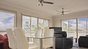 A spacious living room with a glass-top coffee table, white and black leather sofas, and a flat-screen TV. - Hampton Court Apartments (Clayfield)