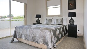 A bedroom with a large bed, two bedside tables, a window, and a view of the outside. - Hampton Court Apartments (Clayfield)