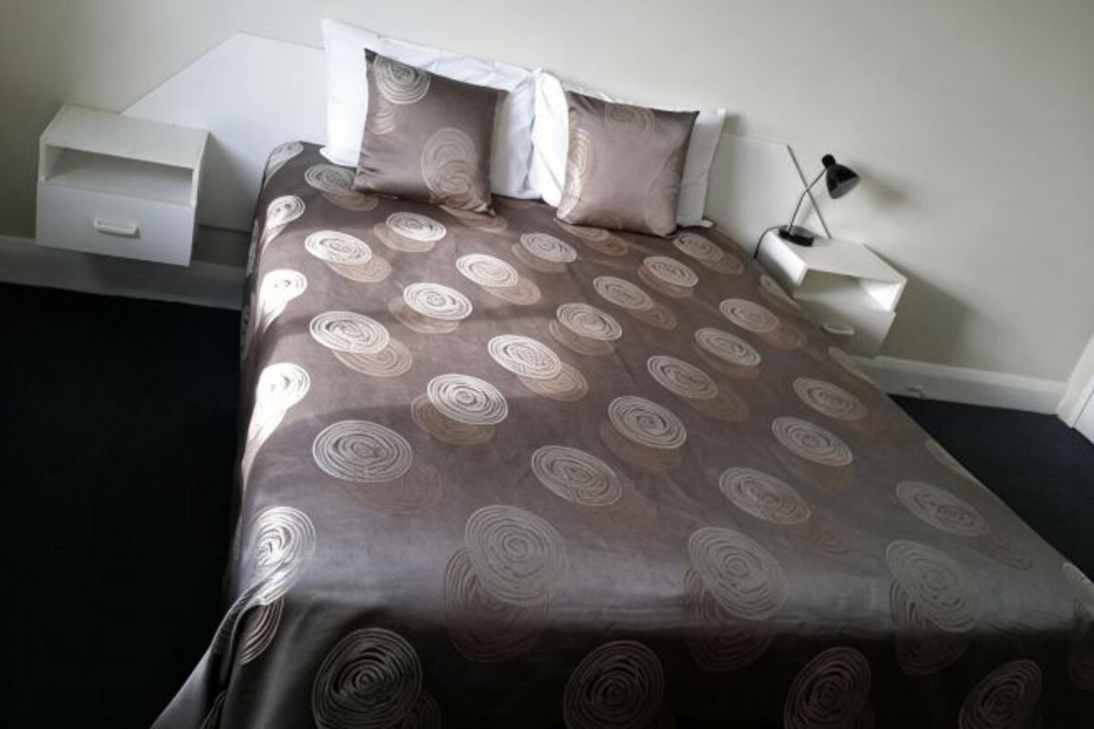 Una cama bien tendida con una colcha estampada, una mesita de noche con una lámpara y otra mesita con una lámpara.
