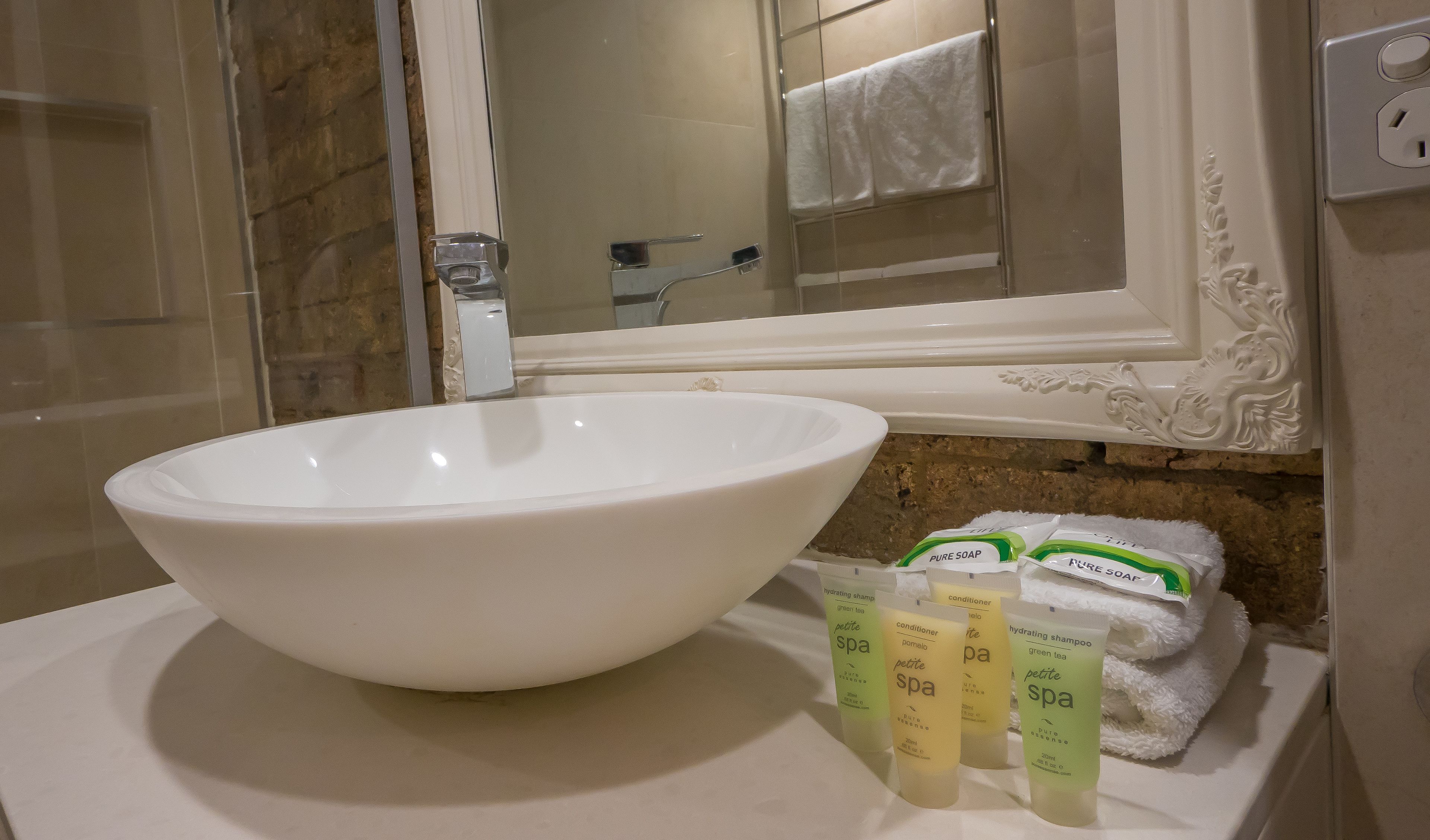 A bathroom with a large white sink, a mirror, and a towel rack. There are three bottles of 'Petite Spa' products on the counter.