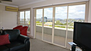 A living room with a large sliding glass door leading to a balcony, a black leather sofa, a red chair, and a flat-screen TV on a glass stand. - Hampton Court Apartments (Clayfield)
