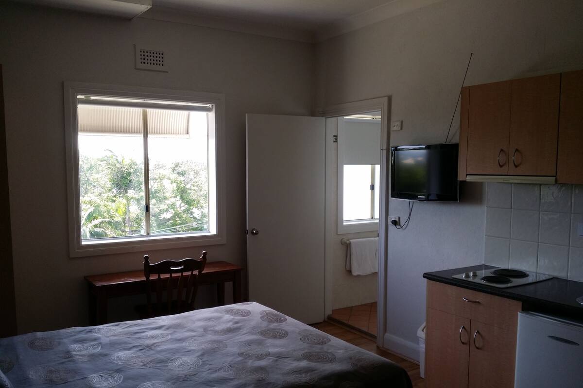 Une petite pièce bien éclairée, avec un lit, une table, un téléviseur fixé au mur et une fenêtre donnant sur un paysage verdoyant.